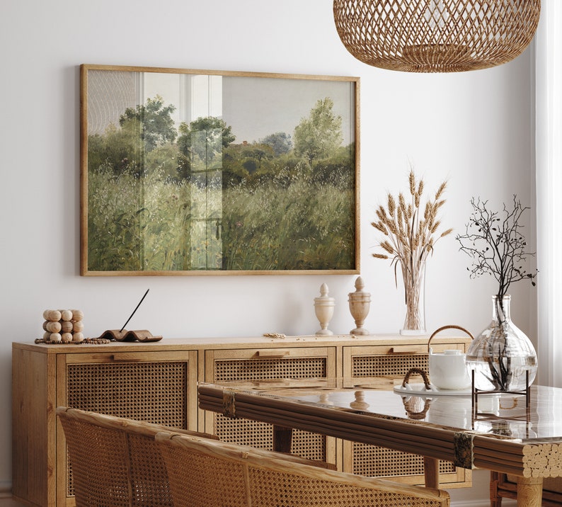 May include: A framed landscape painting in a wooden frame hangs above a wooden cabinet. The painting shows a field of tall grass and trees in green and grey tones. A woven pendant light hangs from the ceiling. The room is decorated with natural elements.