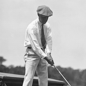 Op de afbeelding: Een man in een wit overhemd, een gestreepte das en een pet houdt een golfclub vast. Hij staat op een golfbaan.