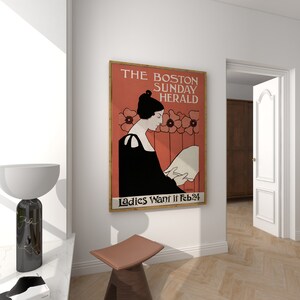 May include: A framed vintage poster for The Boston Sunday Herald, featuring a woman in black reading. The poster has a red background with floral accents and the text "Ladies Want it Feb 24". The poster is displayed on a white wall.