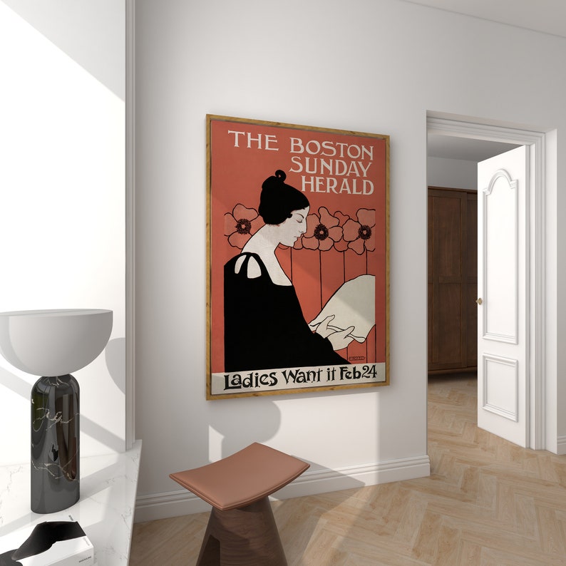 May include: A framed vintage poster for The Boston Sunday Herald, featuring a woman in black reading. The poster has a red background with floral accents and the text "Ladies Want it Feb 24". The poster is displayed on a white wall.