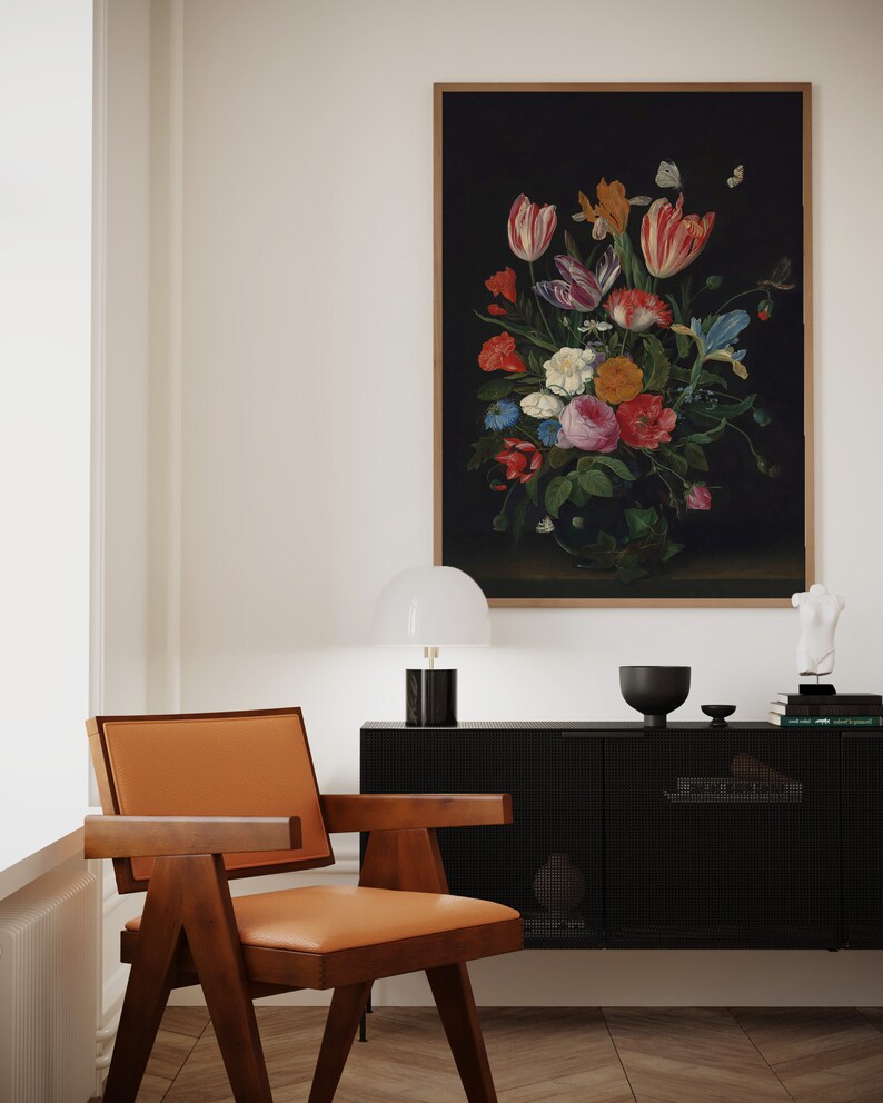 K&ouml;nnte beinhalten: Ein gerahmtes Stillleben mit Blumen vor dunklem Hintergrund h&auml;ngt &uuml;ber einem schwarzen Schrank. Das Gem&auml;lde zeigt einen Strau&szlig; bunter Blumen, darunter Tulpen und Rosen. Ein Stuhl im Mid-Century-Modern-Stil mit braunem Holzrahmen und hellbraunem Ledersitz steht vor dem Schrank.