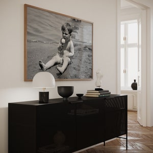 May include: A black metal cabinet with a wire mesh front and two doors. A white lamp with a black base, a black bowl, and a stack of books are on top of the cabinet. A framed black and white photograph of a child eating ice cream on a beach is hanging on the wall behind the cabinet.