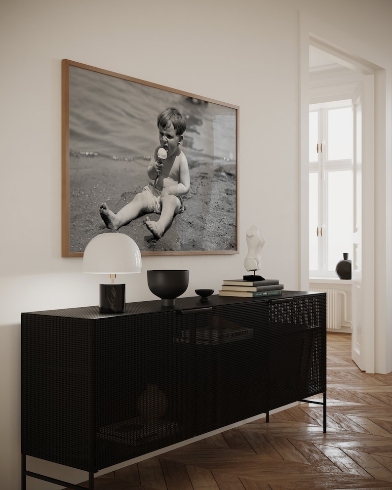 May include: A black metal cabinet with a wire mesh front and two doors. A white lamp with a black base, a black bowl, and a stack of books are on top of the cabinet. A framed black and white photograph of a child eating ice cream on a beach is hanging on the wall behind the cabinet.