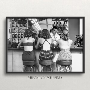 Op de afbeelding: Zwart-witfoto van drie vrouwen die aan een bar zitten in badpakken. Een man staat achter de bar. De vrouwen kijken naar de man. De bar is bedekt met flessen en fruit.