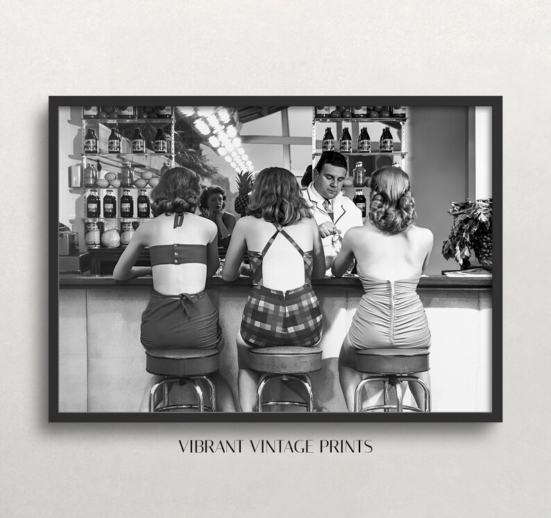 Op de afbeelding: Zwart-witfoto van drie vrouwen die aan een bar zitten in badpakken. Een man staat achter de bar. De vrouwen kijken naar de man. De bar is bedekt met flessen en fruit.