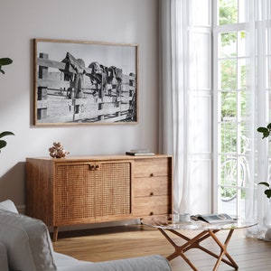 K&ouml;nnte beinhalten: Ein Wohnzimmer im Stil der Mid-Century Modern mit einem Schrank aus Holz mit einer geflochtenen Rattan-T&uuml;r, einem Glas-Couchtisch und einem Schwarzwei&szlig;foto von Pferden, das an der Wand h&auml;ngt.