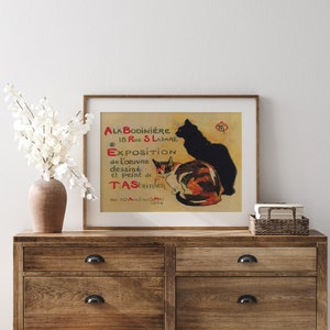 May include: A framed vintage poster featuring two cats, one black and one calico, with text in French. The poster is displayed on a wooden dresser with a vase of white flowers. The poster's text includes "ALA BODINIERE" and "EXPOSITION".
