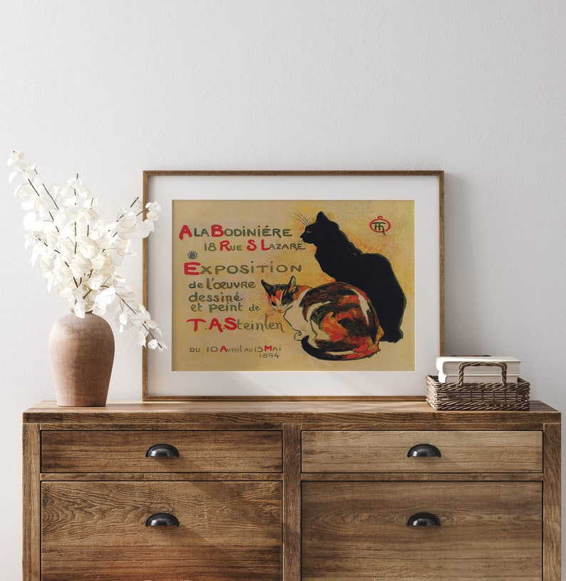 May include: A framed vintage poster featuring two cats, one black and one calico, with text in French. The poster is displayed on a wooden dresser with a vase of white flowers. The poster's text includes "ALA BODINIERE" and "EXPOSITION".