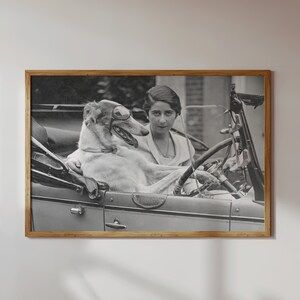 May include: Black and white framed print of a woman and a dog in a vintage car. The dog, a Borzoi, sits in the passenger seat. The woman is behind the steering wheel. The image is in a wooden frame.