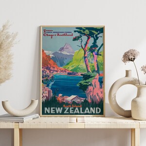 May include: Vintage travel poster advertising scenic views of New Zealand's South Island. The poster features a vibrant landscape with mountains, a lake, and trees. The text reads "Famous Lakes Mountains Fiords Otago & Southland South Island NEW ZEALAND".