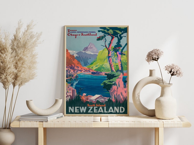 May include: Vintage travel poster advertising scenic views of New Zealand's South Island. The poster features a vibrant landscape with mountains, a lake, and trees. The text reads "Famous Lakes Mountains Fiords Otago & Southland South Island NEW ZEALAND".