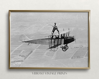 Op de afbeelding: Een zwart-witte vintage foto van een man die op de vleugel van een dubbeldekker in de lucht staat. De man houdt een stok in zijn hand en kijkt naar de camera. De dubbeldekker vliegt over een veld met gewassen.