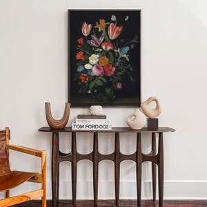 K&ouml;nnte beinhalten: Ein gerahmtes Blumenstillleben-Gem&auml;lde h&auml;ngt &uuml;ber einer Konsole aus dunklem Holz. Auf dem Tisch stehen dekorative Vasen, B&uuml;cher und eine kleine Skulptur. Links ist ein Lederstuhl zu sehen. Das Gem&auml;lde zeigt einen Strau&szlig; bunter Blumen.