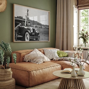 Op de afbeelding: Een bruine leren hoekbank met een wit en zwart gepatroneerd kussen, een groen kussen en een witte deken. Een houten salontafel met een boek en twee witte vazen met bloemen staat voor de bank. Een zwart-wit foto van een vintage auto hangt aan de muur achter de bank.