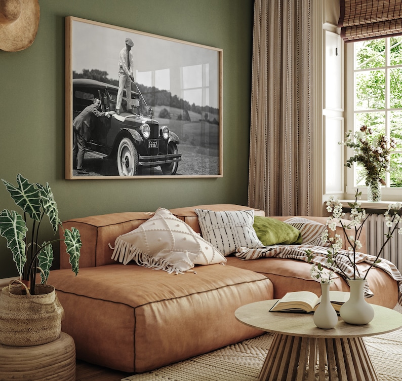 Op de afbeelding: Een bruine leren hoekbank met een wit en zwart gepatroneerd kussen, een groen kussen en een witte deken. Een houten salontafel met een boek en twee witte vazen met bloemen staat voor de bank. Een zwart-wit foto van een vintage auto hangt aan de muur achter de bank.
