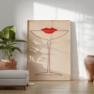 May include: A rectangular framed wall art print featuring a minimalist dark red line drawing of a martini glass centred on a light beige background. A pair of bold, solid red lips is positioned directly above the rim of the glass.
