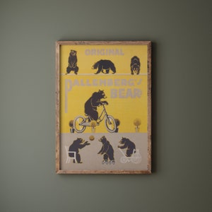 Op de afbeelding: Een ingelijste print met een gele achtergrond en zwarte berenillustraties. De print leest "ORIGINAL DALLENBERG'S BEAR" en toont beren op een koord, fietsend en spelend met een bal. Het frame is gemaakt van lichtbruin hout.