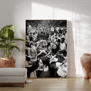 Puede incluir: Fotograf&iacute;a en blanco y negro de una multitud de personas sentadas en la hierba. Un cartel en el centro de la imagen dice "Eve Was Framed".