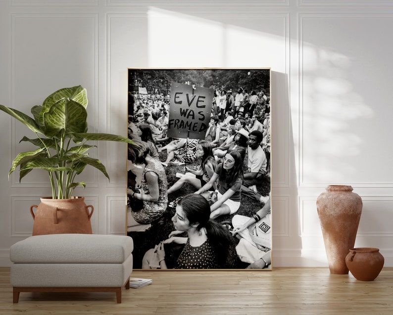 Puede incluir: Fotograf&iacute;a en blanco y negro de una multitud de personas sentadas en la hierba. Un cartel en el centro de la imagen dice "Eve Was Framed".