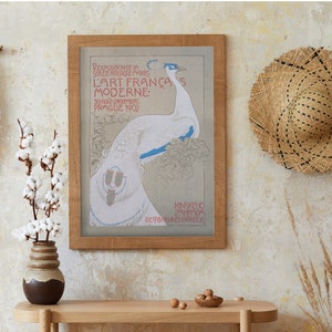 Op de afbeelding: Een ingelijste vintage poster met een pauwenillustratie, met tekst in het Frans. De poster staat in een licht houten lijst, tentoongesteld op een licht houten tafel. Een strohoed hangt aan de muur en een vaas met katoenstelen staat op de tafel.