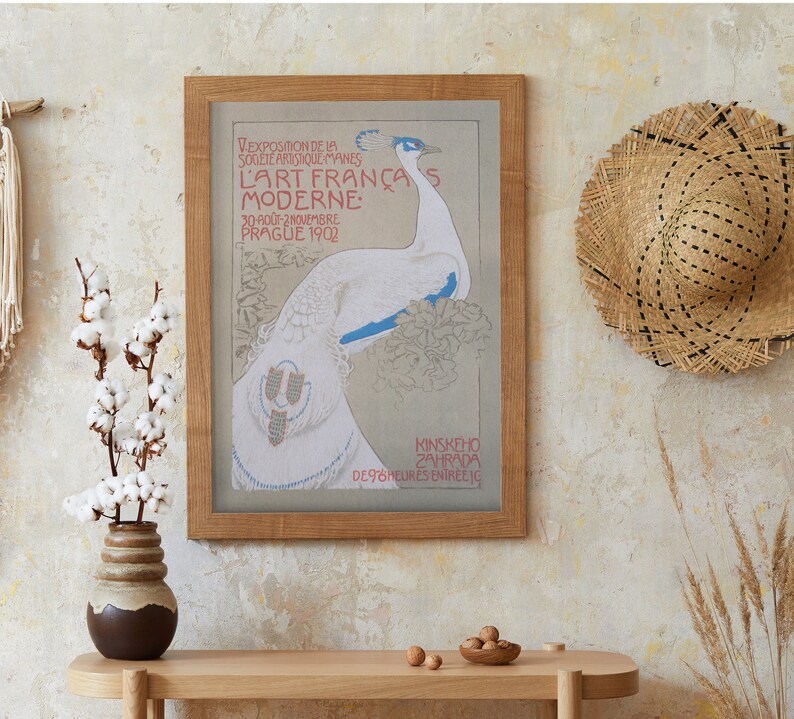 Op de afbeelding: Een ingelijste vintage poster met een pauwenillustratie, met tekst in het Frans. De poster staat in een licht houten lijst, tentoongesteld op een licht houten tafel. Een strohoed hangt aan de muur en een vaas met katoenstelen staat op de tafel.