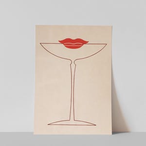 May include: A rectangular art print on a light beige background featuring a minimalist dark red line drawing of a stemmed cocktail glass. Positioned at the top of the glass is a solid red graphic illustration of a pair of lips.