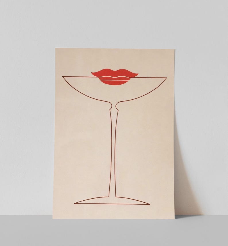 May include: A rectangular art print on a light beige background featuring a minimalist dark red line drawing of a stemmed cocktail glass. Positioned at the top of the glass is a solid red graphic illustration of a pair of lips.