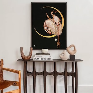 Op de afbeelding: Een ingelijste kunstdruk met een figuur in een wit kledingstuk met patronen, zittend op een gouden halve maan tegen een zwarte achtergrond. Daaronder staat op een donkerhouten consoletafel een bruine U-vormige vaas, een wit object en een Tom Ford 002 boek.