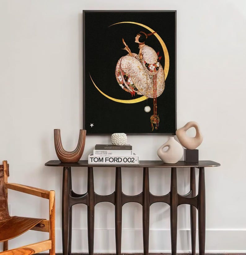 Op de afbeelding: Een ingelijste kunstdruk met een figuur in een wit kledingstuk met patronen, zittend op een gouden halve maan tegen een zwarte achtergrond. Daaronder staat op een donkerhouten consoletafel een bruine U-vormige vaas, een wit object en een Tom Ford 002 boek.
