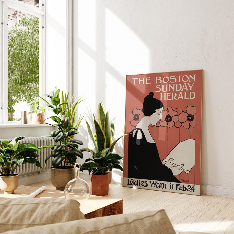 May include: A framed vintage poster for The Boston Sunday Herald, featuring a woman in black attire reading. The poster has a red background with floral accents and the text "Ladies Want it Feb 24". Several potted plants are in the foreground.