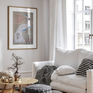 Op de afbeelding: Lichte woonkamer met een ingelijste vintage poster, een witte bank met kussens en een gestreepte deken, een houten salontafel en een grijze poef. De kamer heeft een lichte en luchtige uitstraling.