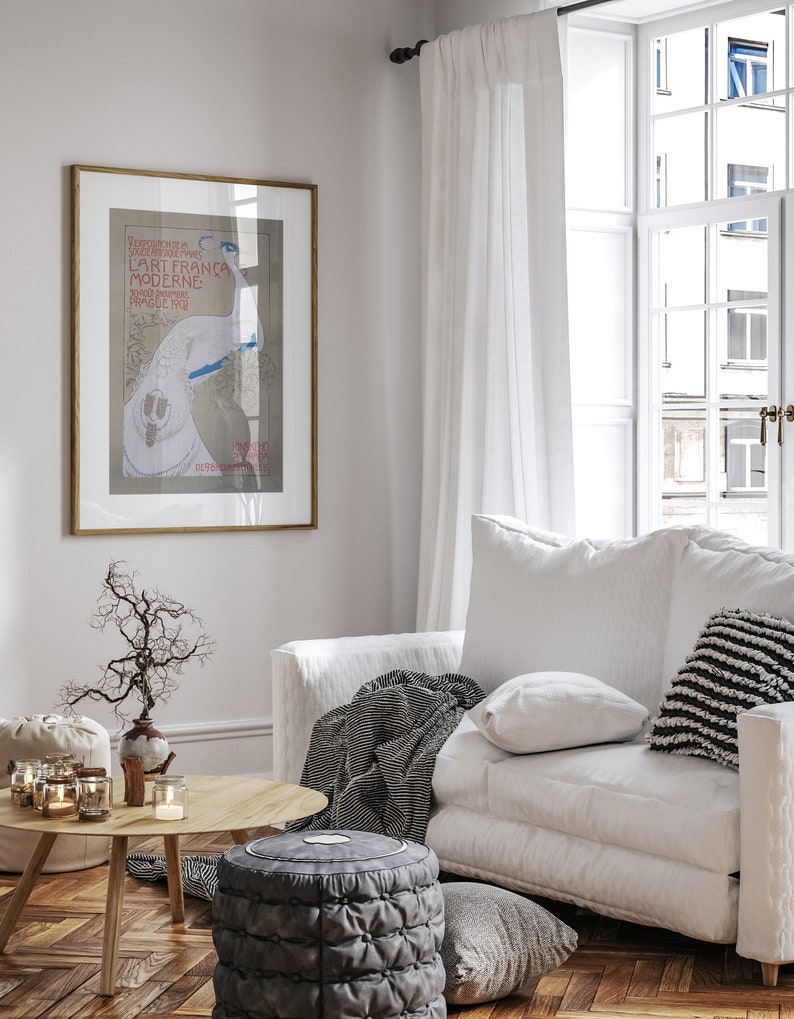 Op de afbeelding: Lichte woonkamer met een ingelijste vintage poster, een witte bank met kussens en een gestreepte deken, een houten salontafel en een grijze poef. De kamer heeft een lichte en luchtige uitstraling.