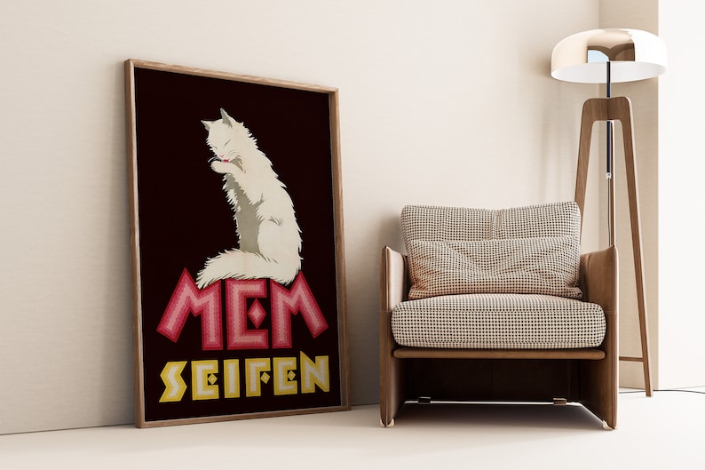 May include: Framed vintage poster featuring a white cat grooming itself, set against a dark maroon background. The poster has the words "MEM SEIFEN" in red and yellow. The poster is displayed next to a wooden chair.
