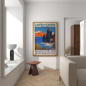 May include: A framed vintage travel poster for Lake of Garda, Italy, featuring a colorful landscape with mountains, water, and trees. The poster has the text "LAKE OF GARDA" and "GARDONE RIV. AND FASANO". The poster is in a room with a window and a wooden stool.