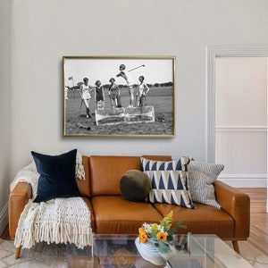 May include: Black and white photograph of five women playing golf on a sunny day. The women are wearing dresses and are standing on a golf course. The photo is framed in a gold frame and is hanging on a wall.