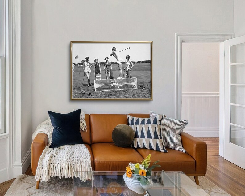 May include: Black and white photograph of five women playing golf on a sunny day. The women are wearing dresses and are standing on a golf course. The photo is framed in a gold frame and is hanging on a wall.
