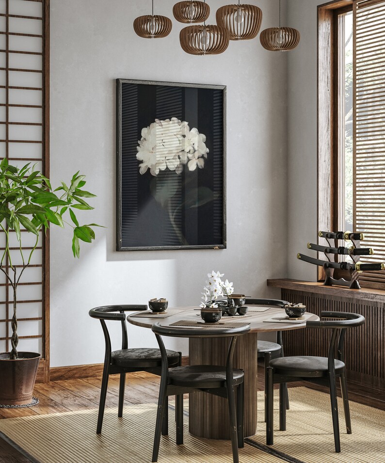 May include: A round wooden dining table with four black chairs. The table has a white flower arrangement in the center and a black framed print of a white flower on a dark background hanging on the wall behind it.