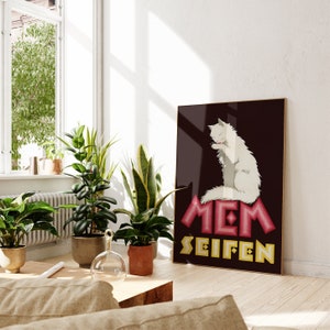 May include: A framed poster features a white cat grooming itself on a pink and yellow geometric design. The poster has the German words "MEM SEIFEN" in a stylized font against a dark brown background. The poster is displayed in a bright room with plants.