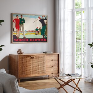May include: A framed vintage golf poster with the text "North Berwick" in red. The poster depicts golfers in period attire. The poster is displayed above a wooden cabinet with a woven door and three drawers. A glass-topped coffee table and white sofa are in the foreground.