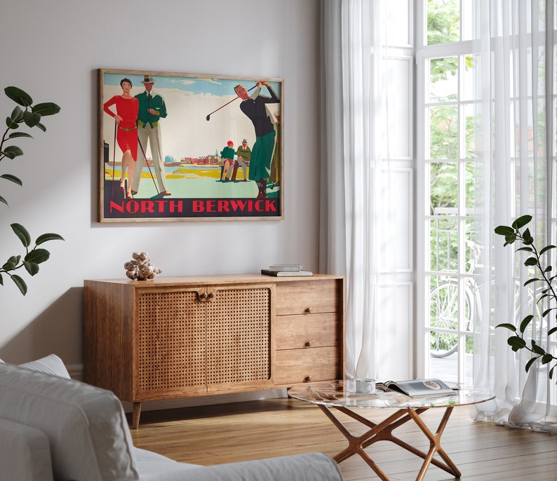 May include: A framed vintage golf poster with the text "North Berwick" in red. The poster depicts golfers in period attire. The poster is displayed above a wooden cabinet with a woven door and three drawers. A glass-topped coffee table and white sofa are in the foreground.