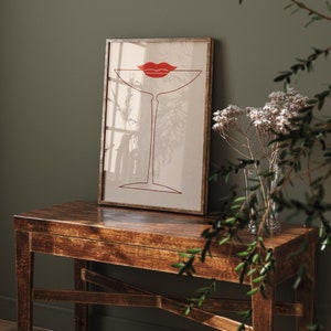 May include: A framed wall art print featuring a minimalist red line drawing of a martini glass with a pair of red lips positioned at the rim. The artwork is set against a light beige background, enclosed in a thin, dark-toned wooden frame.