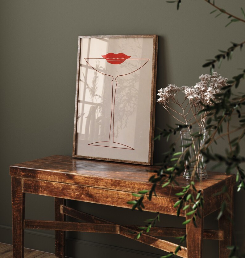 May include: A framed wall art print featuring a minimalist red line drawing of a martini glass with a pair of red lips positioned at the rim. The artwork is set against a light beige background, enclosed in a thin, dark-toned wooden frame.