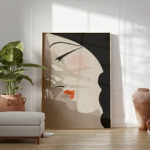 May include: Abstract art print featuring a woman's profile with closed eyes and red lips. The artwork has a neutral color palette with black, beige, and peach tones, framed in gold. The art piece is displayed in a well-lit room with a plant and a neutral-colored ottoman.