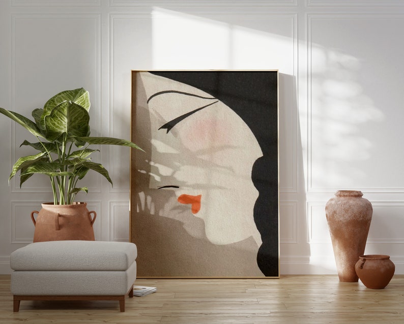 May include: Abstract art print featuring a woman's profile with closed eyes and red lips. The artwork has a neutral color palette with black, beige, and peach tones, framed in gold. The art piece is displayed in a well-lit room with a plant and a neutral-colored ottoman.