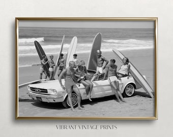 May include: A black and white photographic print featuring a group of people gathered around a vintage convertible car on a sandy beach. Several surfboards are propped up against the vehicle and the car is parked near the ocean waves.