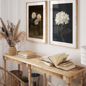 May include: Two framed floral art prints. One shows pink roses in a glass vase, the other a white hydrangea. They hang above a wooden console table with books and decorative items.