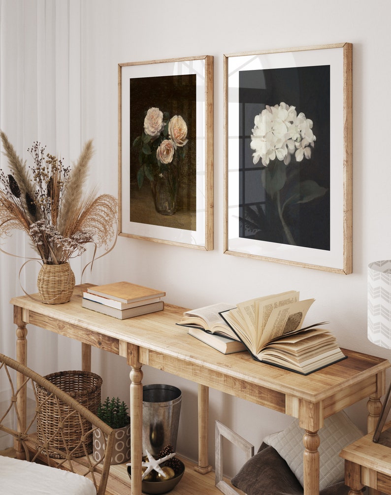 May include: Two framed floral art prints. One shows pink roses in a glass vase, the other a white hydrangea. They hang above a wooden console table with books and decorative items.