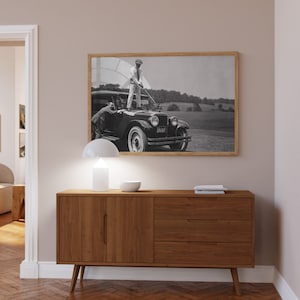 Op de afbeelding: Een vintage zwart-witfoto van een man die op de motorkap van een vintage auto staat. De man houdt een golfclub vast en kijkt naar de camera. De auto heeft een kentekenplaat met de tekst "D1567". De foto is ingelijst in een houten lijst.