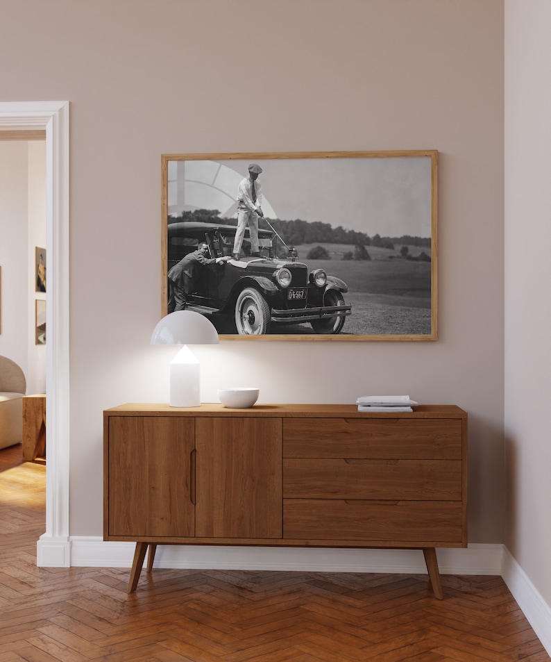 Op de afbeelding: Een vintage zwart-witfoto van een man die op de motorkap van een vintage auto staat. De man houdt een golfclub vast en kijkt naar de camera. De auto heeft een kentekenplaat met de tekst "D1567". De foto is ingelijst in een houten lijst.
