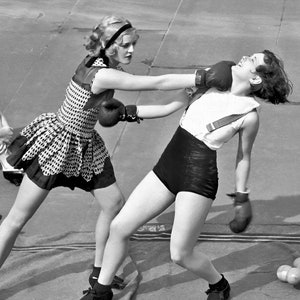 Puede incluir: Fotograf&iacute;a en blanco y negro de dos mujeres boxeando. Una, con un vestido de lunares y guantes de boxeo, lanza un pu&ntilde;etazo. La otra, con una camiseta blanca y pantalones cortos negros, es golpeada. Ambas llevan guantes de boxeo.